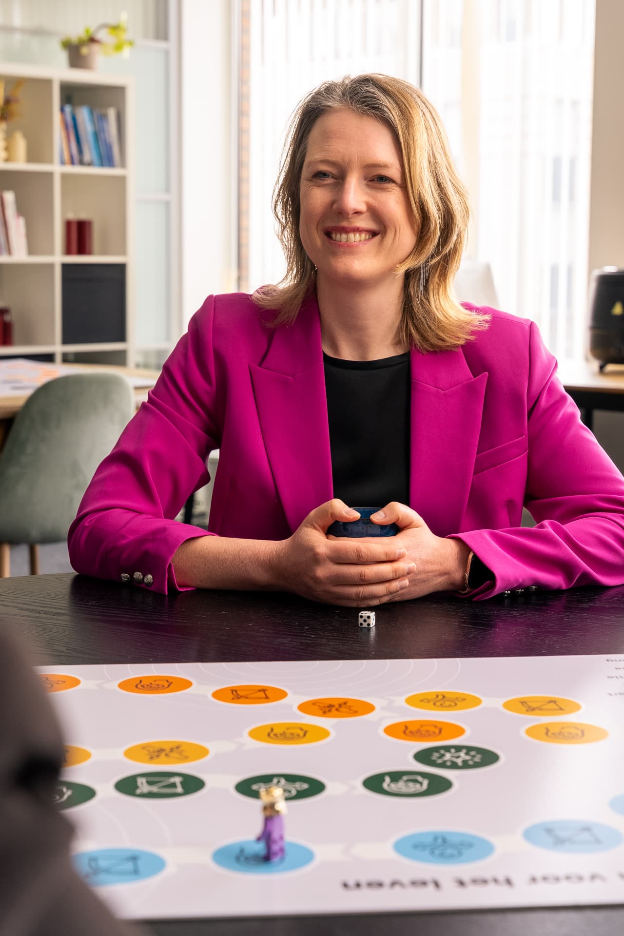Speelster in een fuchsia jasje aan tafel met het bordspel Klanten voor het leven, met witte dobbelsteen en paarse speelfiguur op het speelveld